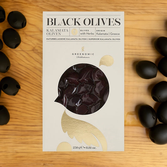 A packaging box for black olives, labeled "BLACK OLIVES," The box has a transparent window showing some of the olives inside. There are scattered black olives on a wooden surface around the box.