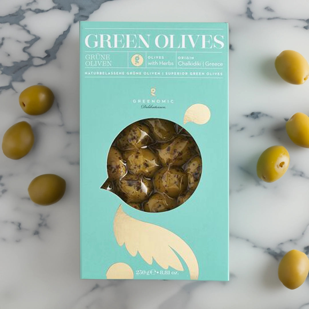 A blue and gold packaging box containing green olives. The box has a transparent window showing some of the olives inside. The olives are seasoned with herbs