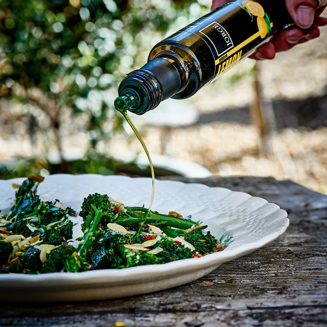 A bottle of lemon oil being drizzled over a tender stem broccoli salad.